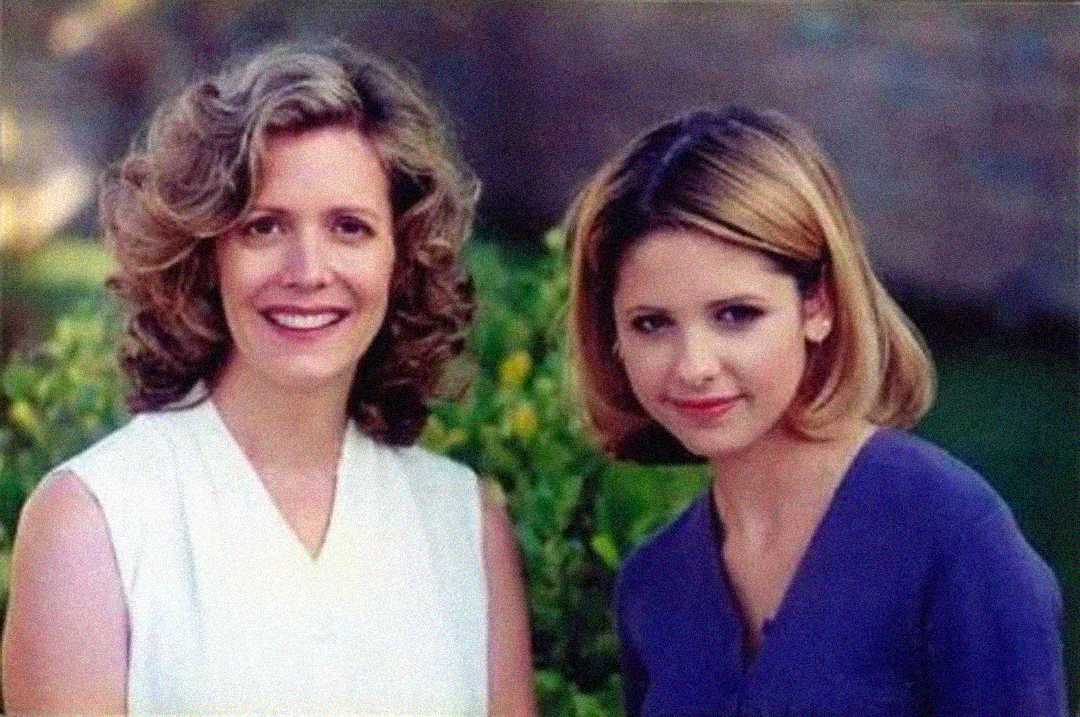 Two women are sitting close together outdoors, smiling at the camera. One has curly, shoulder-length blonde hair and wears a white sleeveless top; the other has straight, chin-length blonde hair and wears a purple shirt. Greenery is in the background.