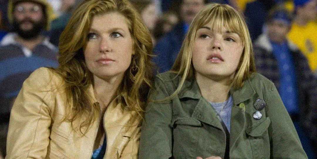 Two women with light brown and blonde hair sit closely together in a stadium, looking ahead with thoughtful expressions. The woman on the left wears a tan jacket; the woman on the right wears an olive jacket with pins.