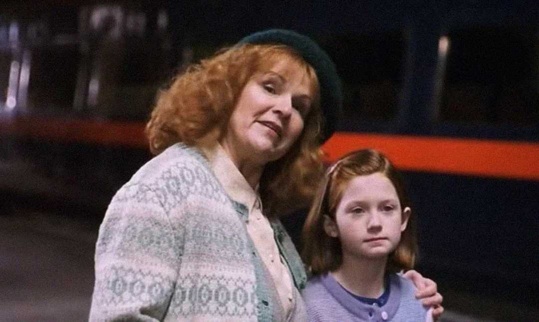 A woman with curly red hair and a green hat stands with her arm around a young girl with straight red hair. They both wear sweaters and stand in front of a dark train with an orange stripe.