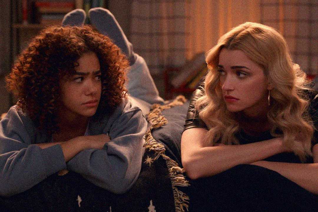 Two young women lie on a bed facing each other, having a serious conversation. One has curly hair and wears a sweatshirt, while the other has long blonde hair and wears a dark top. The room is softly lit.