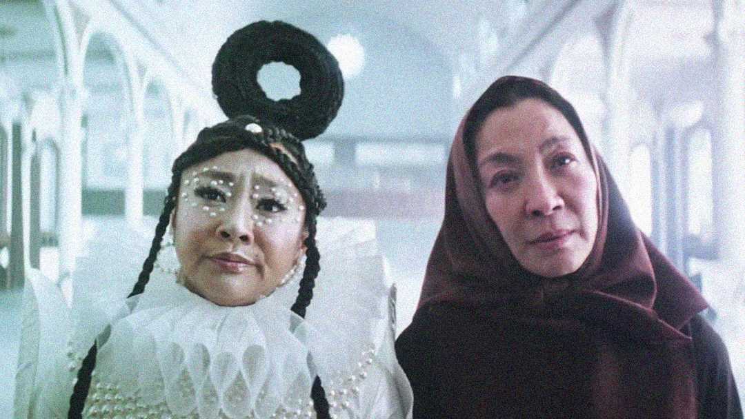 Two women stand side by side in a bright hallway; one wears an elaborate white costume with face decorations and a circular headpiece, while the other wears a simple dark hooded cloak.