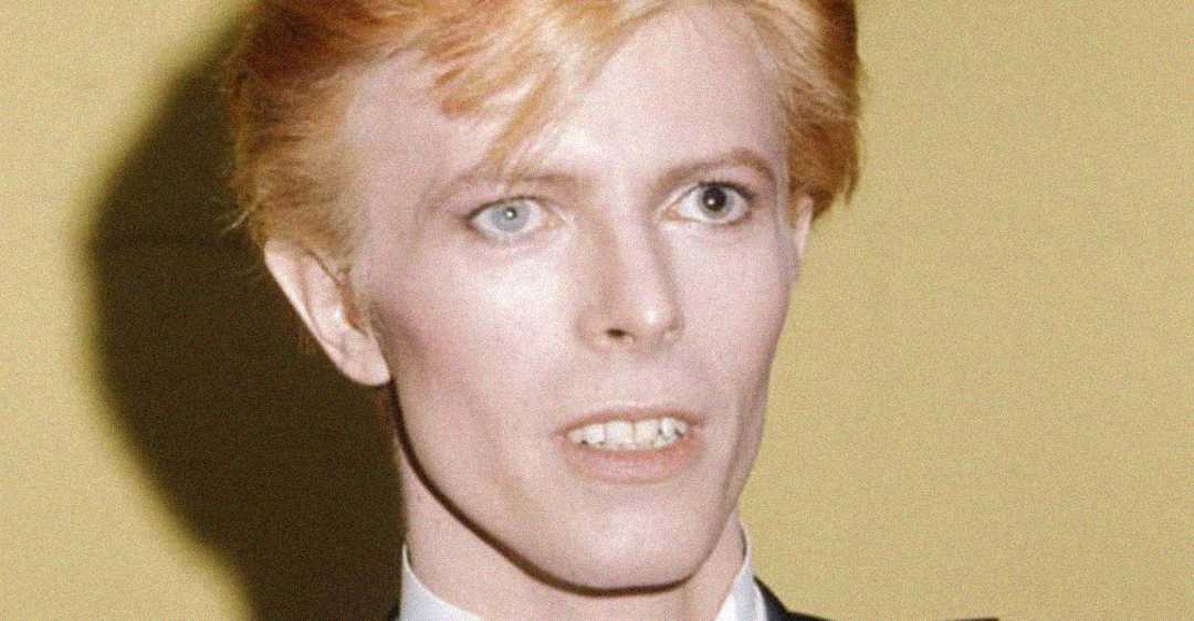 A person with pale skin, light blue eyes, and short strawberry blond hair looks slightly past the camera. They are wearing a dark suit jacket and a white collared shirt, set against a plain yellow background.