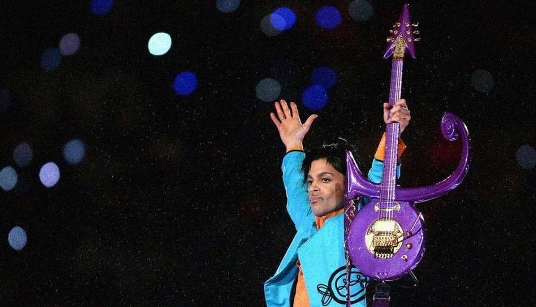 A musician in a bright blue outfit raises a custom purple guitar with one hand on stage, surrounded by glowing lights in the background.