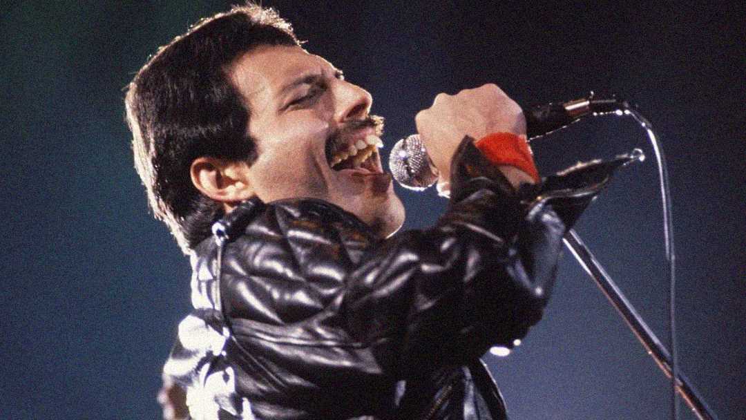 A man with short dark hair and a mustache passionately sings into a microphone on stage, wearing a black leather jacket and red wristband, with dramatic stage lighting in the background.