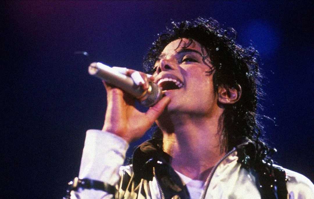 A person with curly black hair passionately sings into a microphone on stage, wearing a white shirt and dark jacket, with dramatic stage lighting in the background.