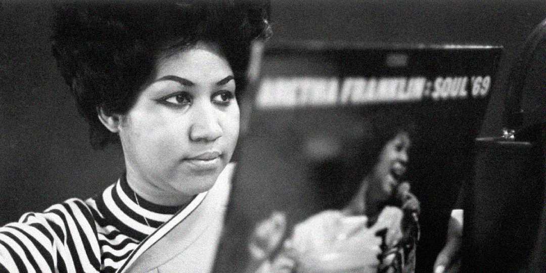 A woman with styled hair and a striped top looks at an album cover featuring her own image and the text “Aretha Franklin - Soul ’69.” The photo is in black and white.