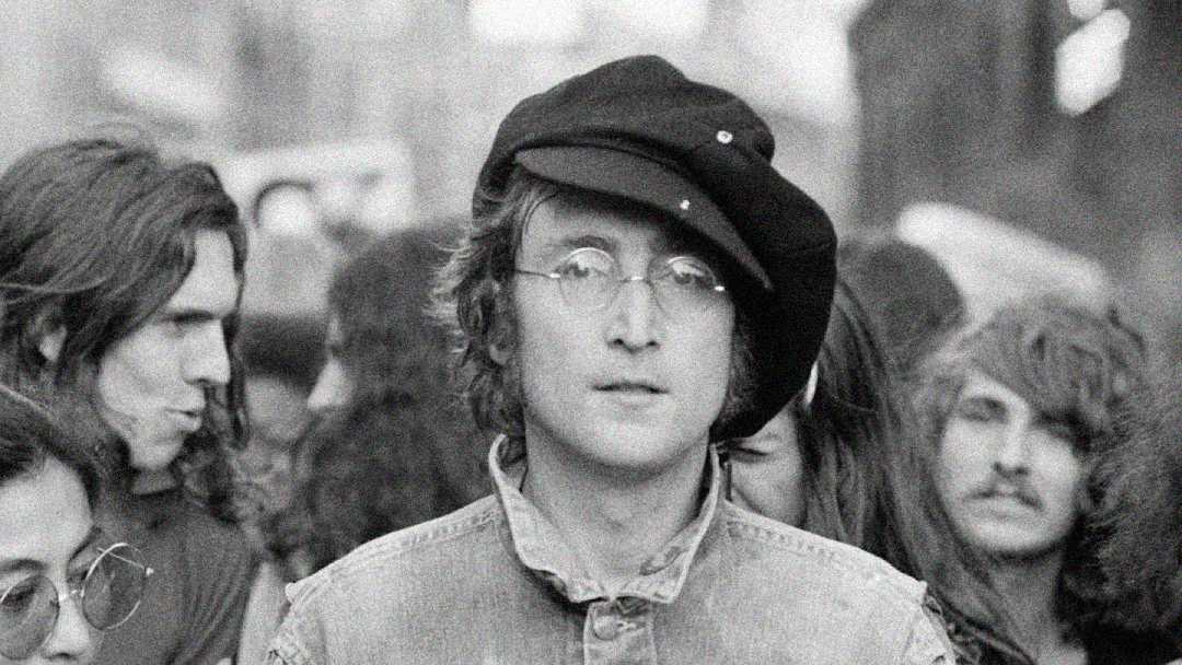 A black and white photo of a man wearing round glasses, a denim jacket, and a tilted beret, standing among a crowd of people with long hair, some looking at the camera, others looking away.