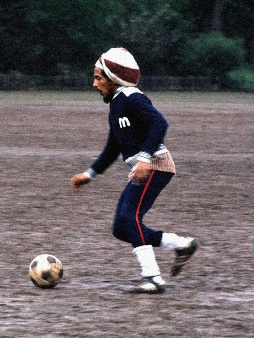 A man wearing a sweater, pants, and a headwrap is playing soccer on a dirt field, mid-stride, focused on controlling the ball. Trees and greenery are visible in the blurred background.