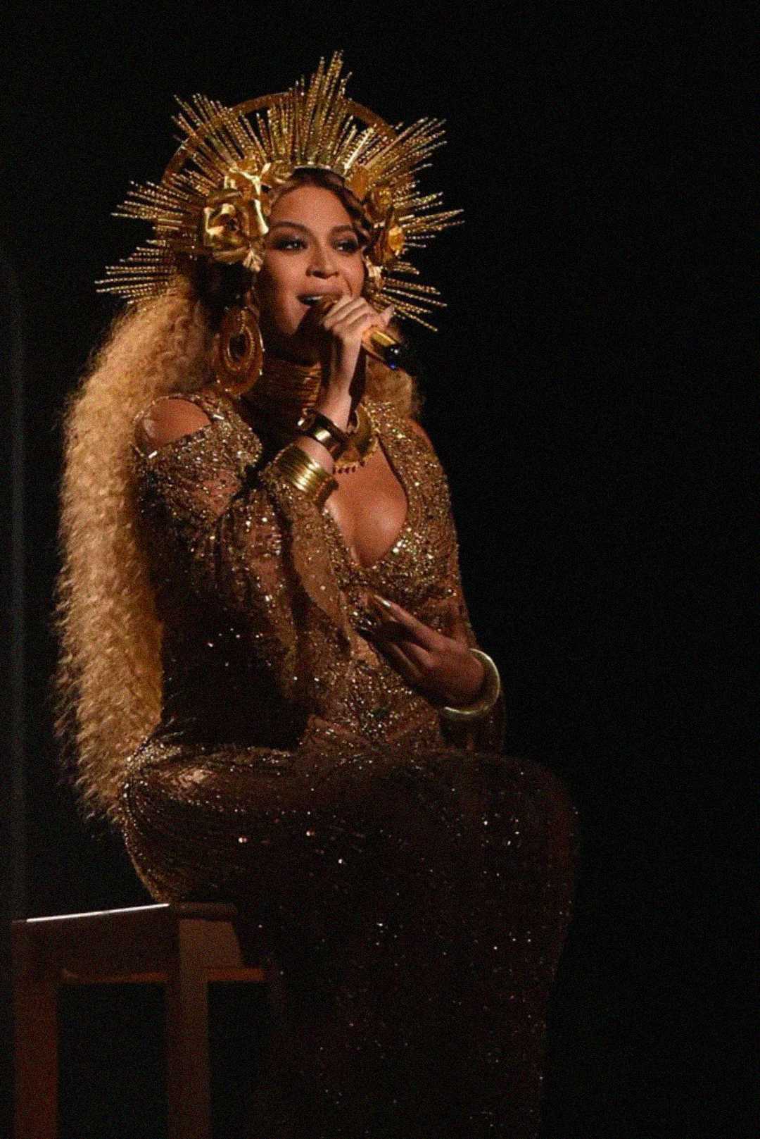 Woman with long curly hair wears a sparkling gold dress and an ornate golden crown with rays, sitting on a stool and singing into a microphone on a dark stage.