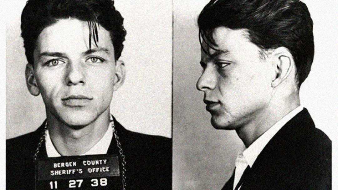 Black-and-white mugshot of a young man with dark hair, wearing a suit. He faces forward in one photo and to the right in the other. He holds a sign reading "Bergen County Sheriff's Office 11 27 38.
