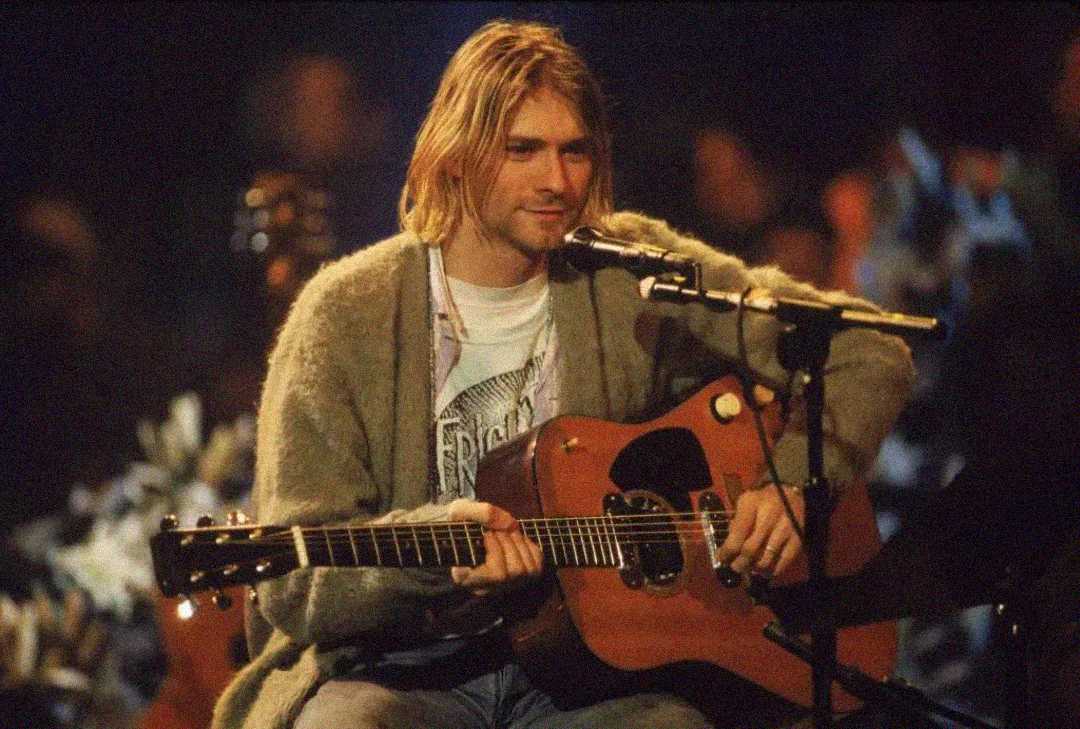 A blond man wearing a cardigan and jeans sits on stage, playing an acoustic guitar and singing into a microphone, with flowers and dim, warm lighting in the background.
