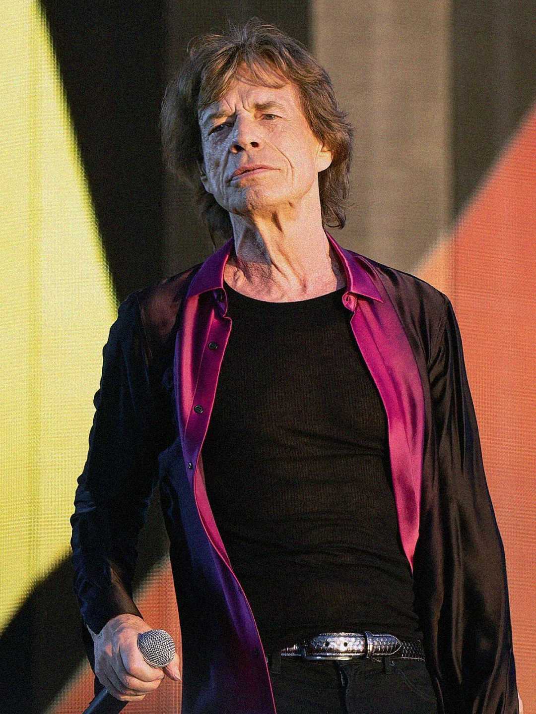 An older man with shoulder-length hair, wearing a black shirt with an open purple and black button-down, stands on stage holding a microphone with a colorful background behind him.