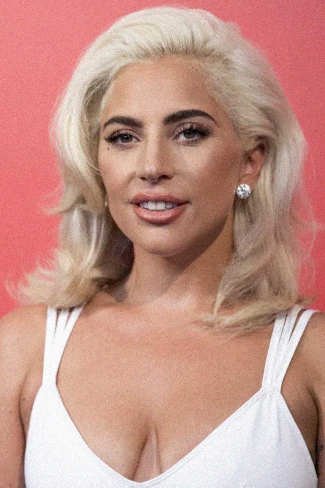A person with platinum blonde hair styled in loose waves, wearing diamond stud earrings and a white top, poses in front of a solid pink background.