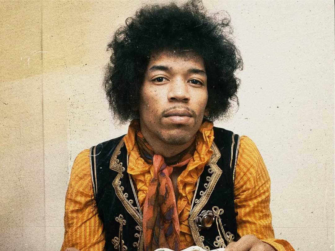A man with curly hair wears a yellow shirt, an ornate black vest, and a red patterned scarf, looking directly at the camera against a plain, light background.