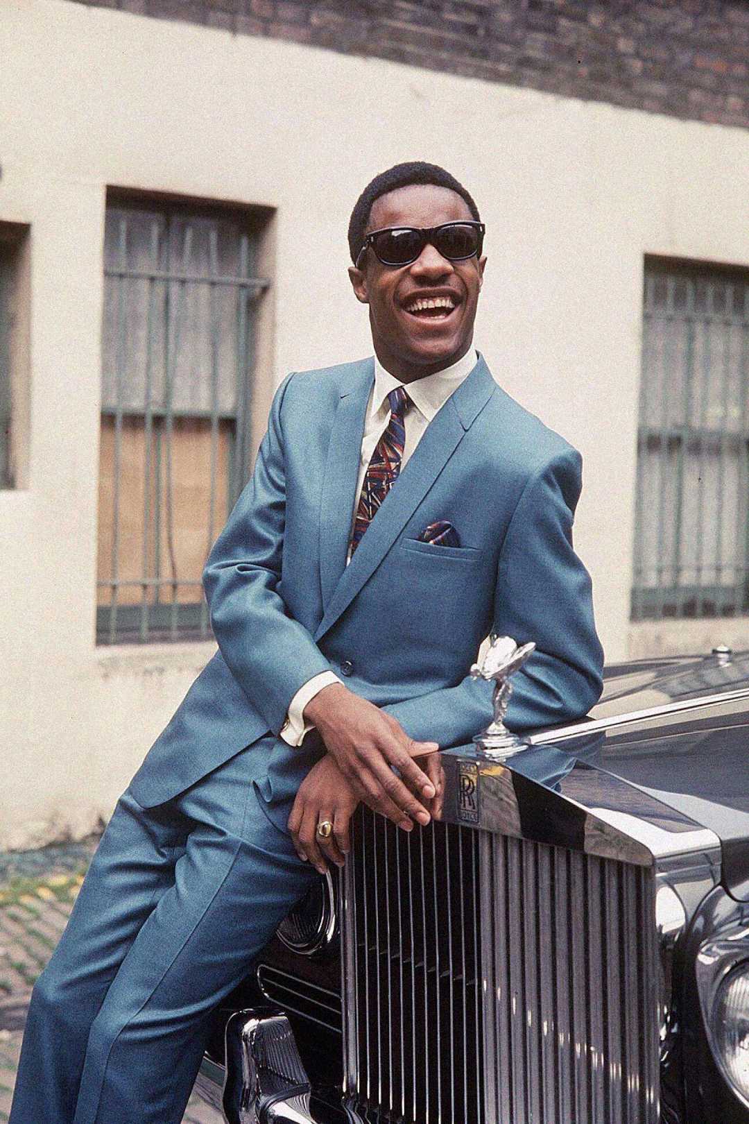 A man wearing sunglasses and a blue suit smiles while leaning against the front of a classic Rolls-Royce car parked on a street, with a building in the background.