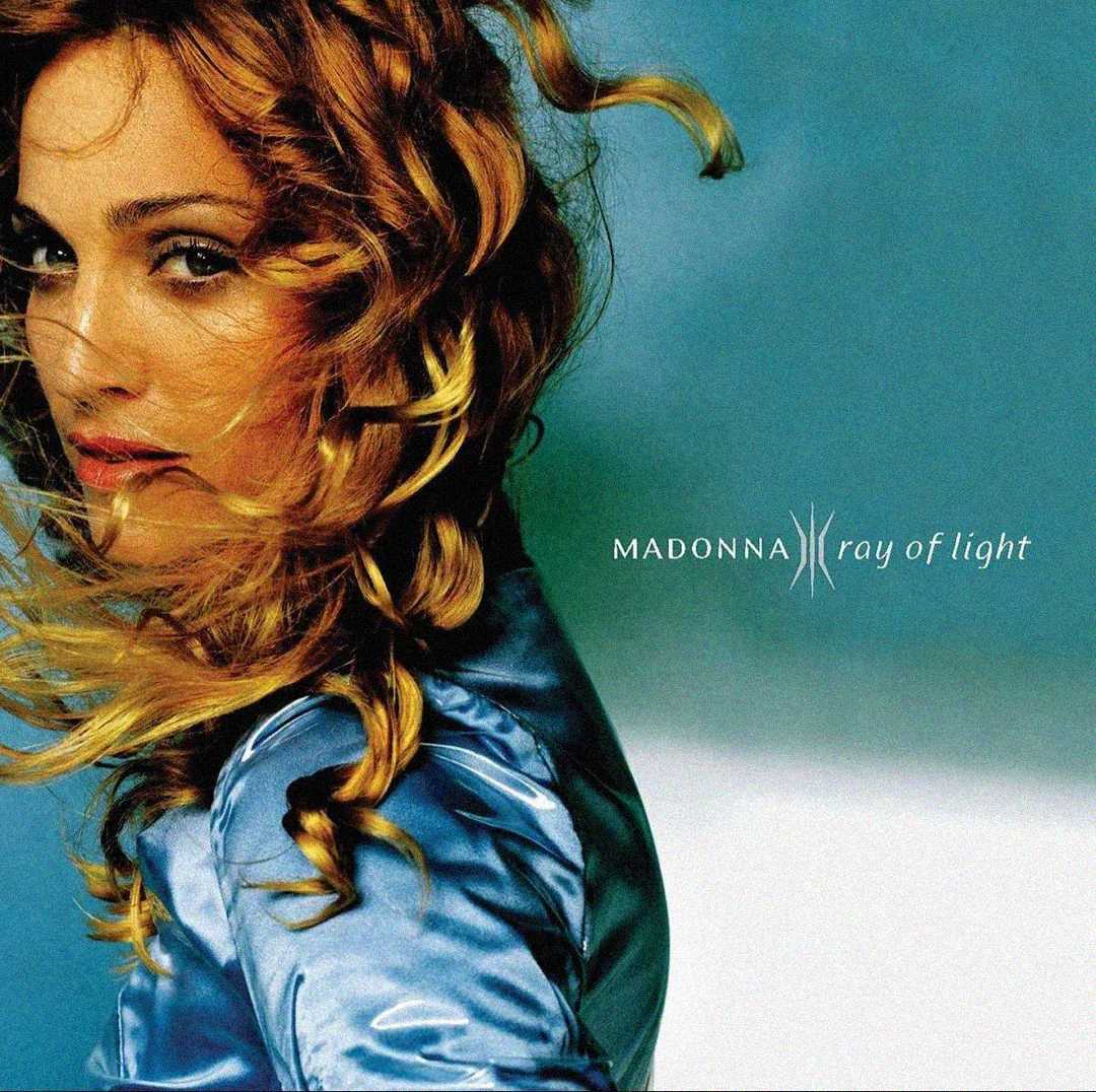 Close-up of a woman with wavy blonde hair wearing a shiny blue top, looking over her shoulder against a blue and green gradient background. Text reads "Madonna Ray of Light.