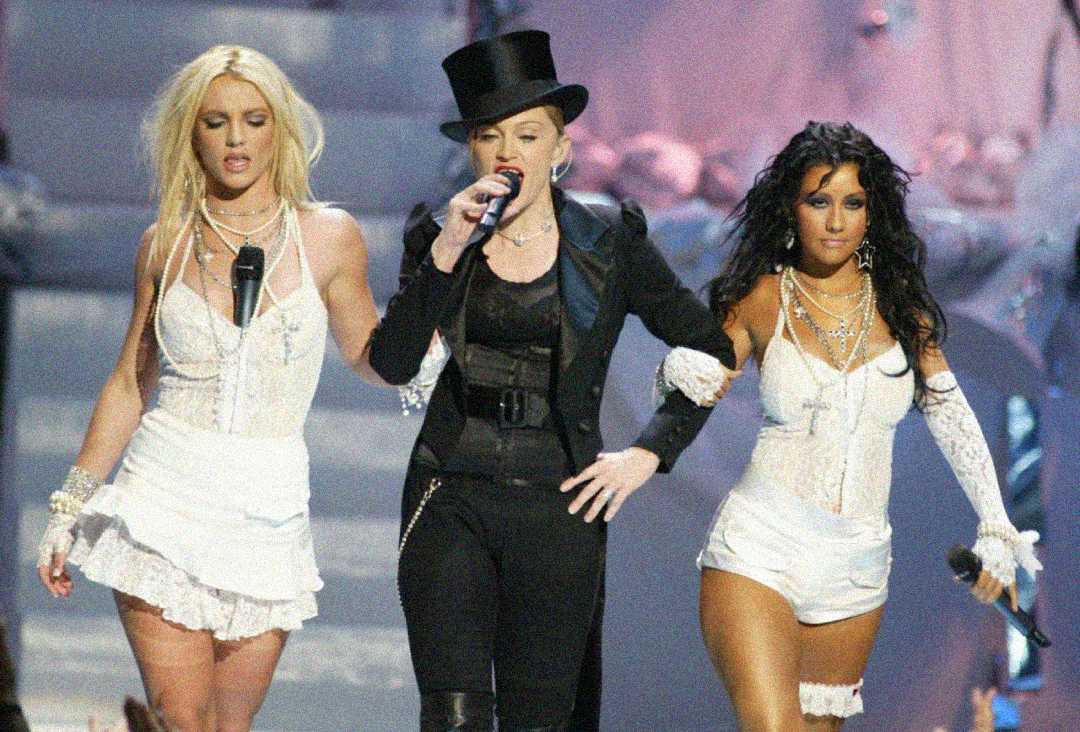 Three women perform on stage in white outfits; the center woman wears a black suit, top hat, and holds a microphone, while the other two wear matching white lingerie-inspired costumes and hold microphones.