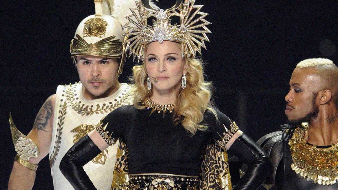A woman in an elaborate gold headdress and black-and-gold costume stands between two men in ornate Roman-inspired outfits during a stage performance.