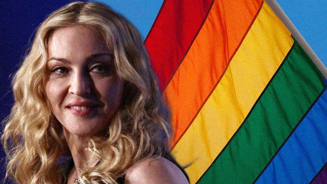 A woman with long, wavy blonde hair smiles in front of a rainbow pride flag against a blue background.