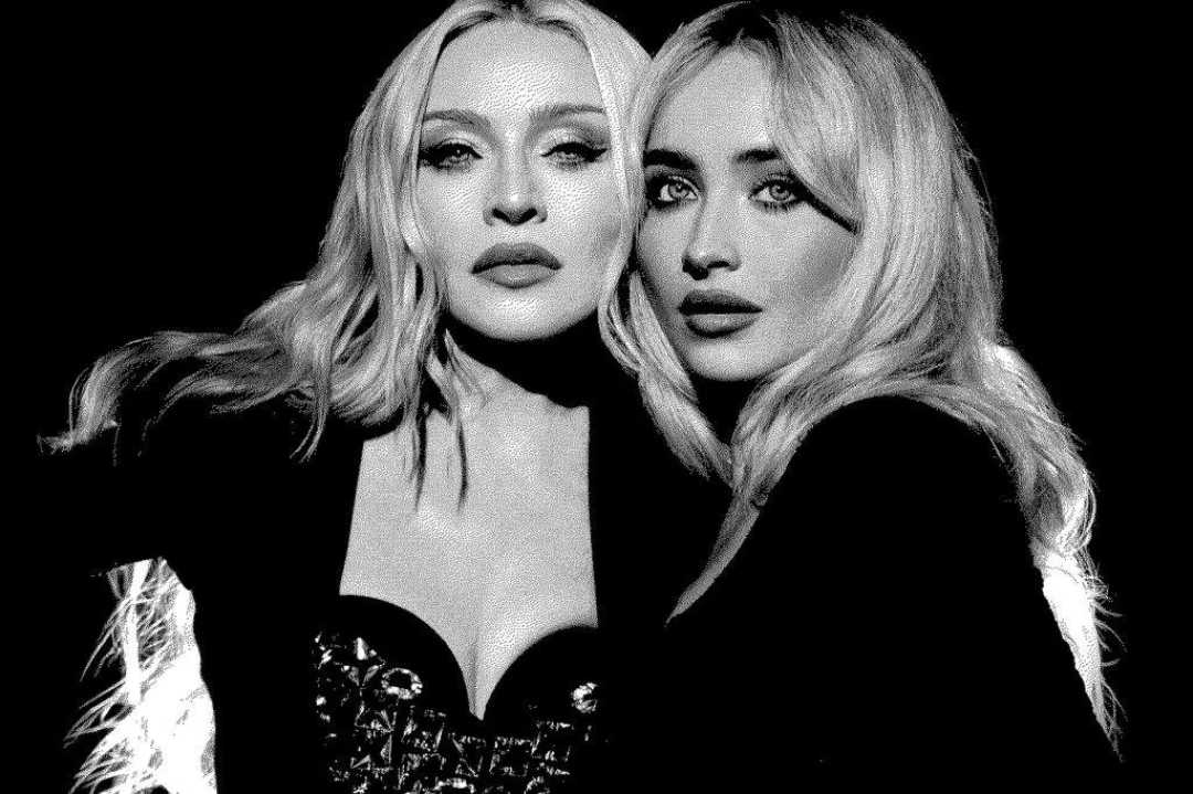 Two women with long blonde hair and dramatic makeup pose closely together in black outfits against a dark background. The photo is in black and white, giving it a classic, high-contrast look.