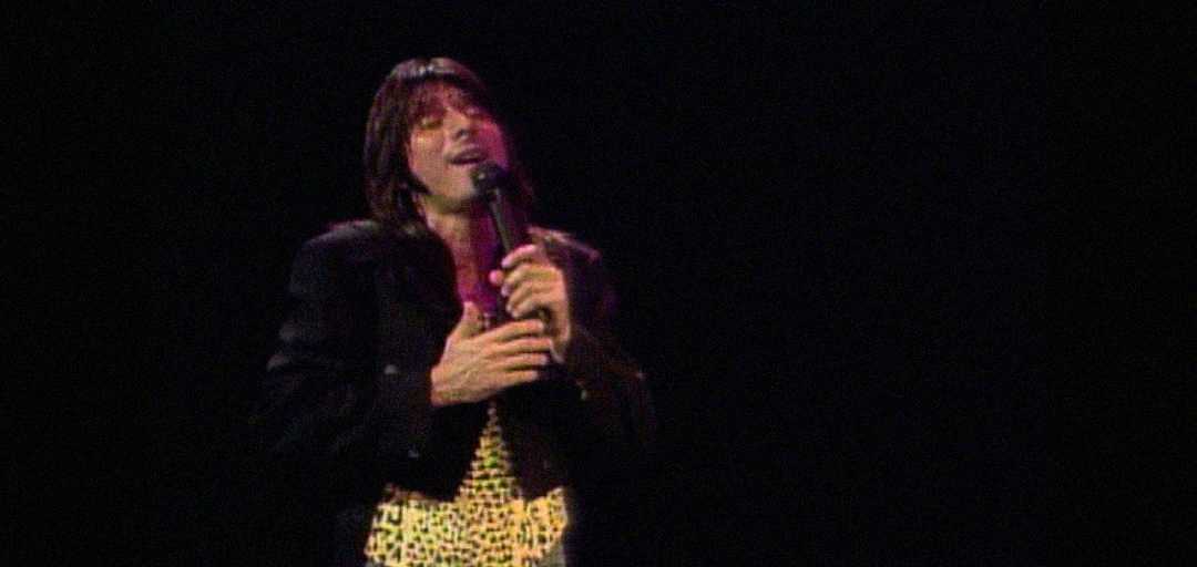 A singer with long dark hair holds a microphone and performs on stage, wearing a black jacket and a yellow leopard-print shirt against a dark background.
