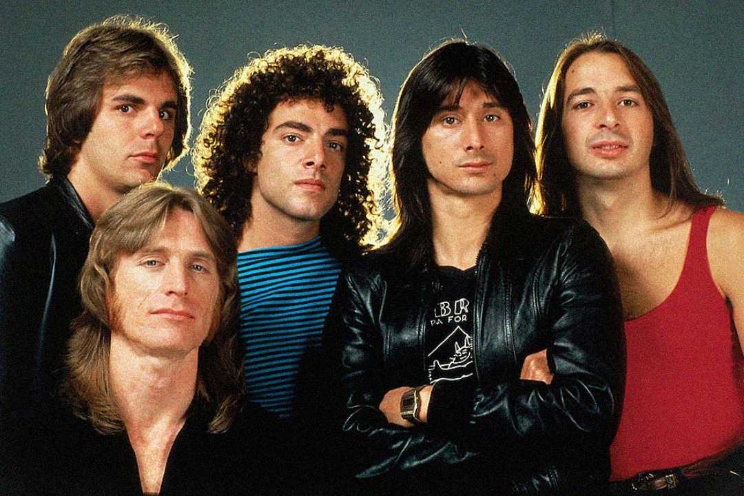 Five men with long hair pose together against a plain background, dressed in casual 1980s-style clothing, including leather jackets, a striped shirt, and a red tank top.