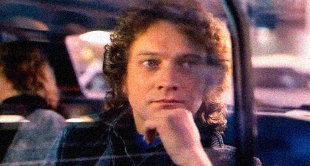 A person with curly hair sits in a car, resting their chin on their hand and gazing thoughtfully out the window. The image is softly focused, with reflections on the glass.
