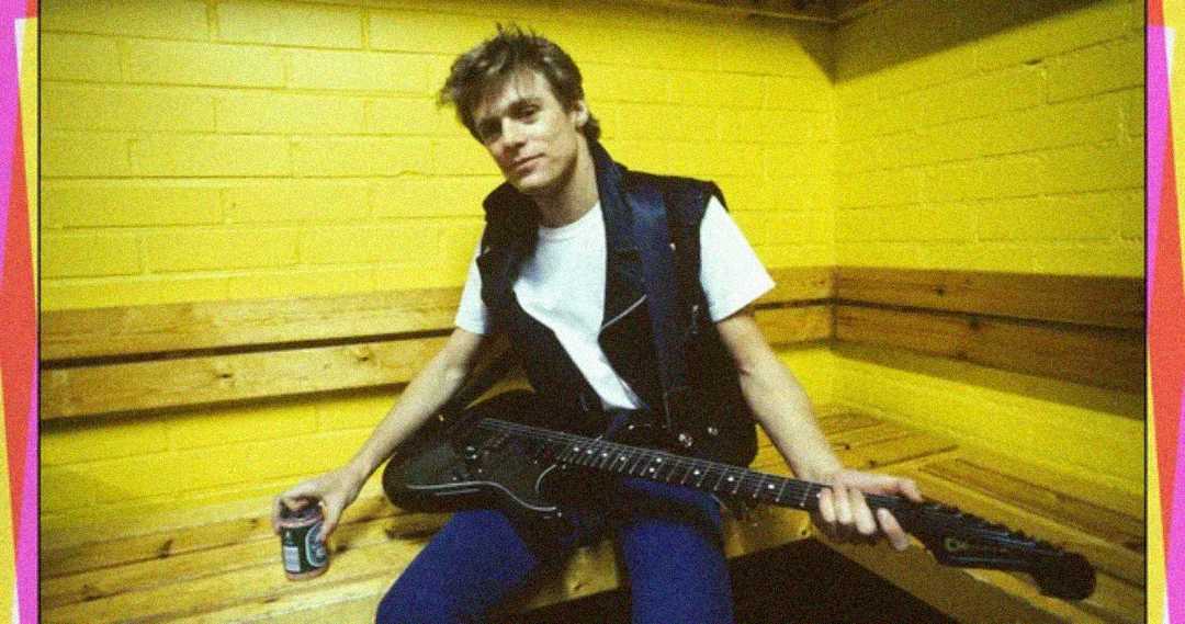 A person in a black leather vest and white shirt sits on a wooden bench against a bright yellow brick wall, holding an electric guitar and a can, looking at the camera with a relaxed expression.