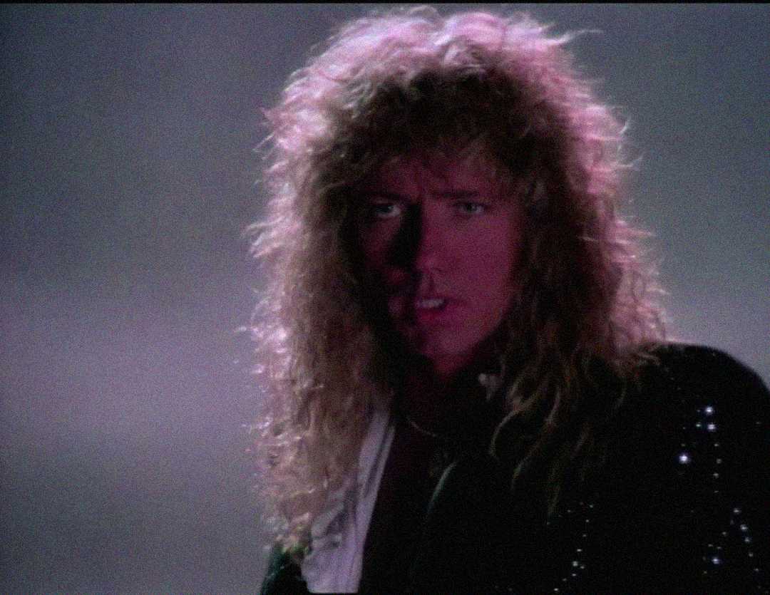 A person with long, curly blond hair and a serious expression looks toward the camera, illuminated by soft, dramatic lighting with a dark, misty background.