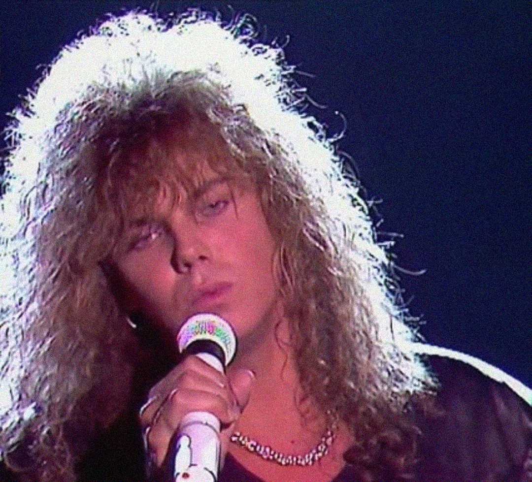 A person with long, curly hair and a chain necklace sings into a microphone under dramatic lighting, giving an intense expression.