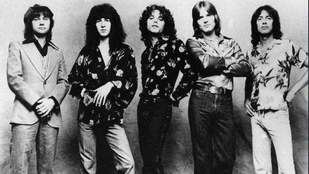 Five men stand in a row against a plain background, wearing 1970s-style clothing including patterned shirts and jeans, with varied hairstyles ranging from curly to straight. The image is in black and white.
