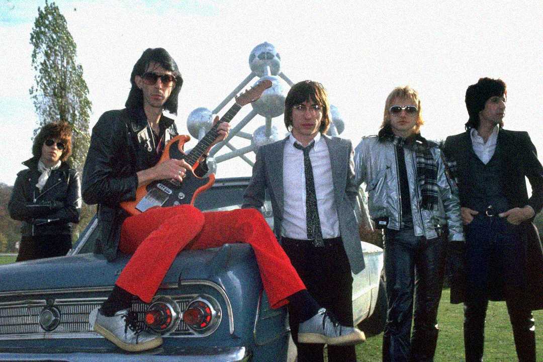 Five men in 1980s-style clothing pose by a vintage blue car. One man sits on the hood with a red electric guitar. The Atomium structure is visible in the background. The mood is casual and cool.