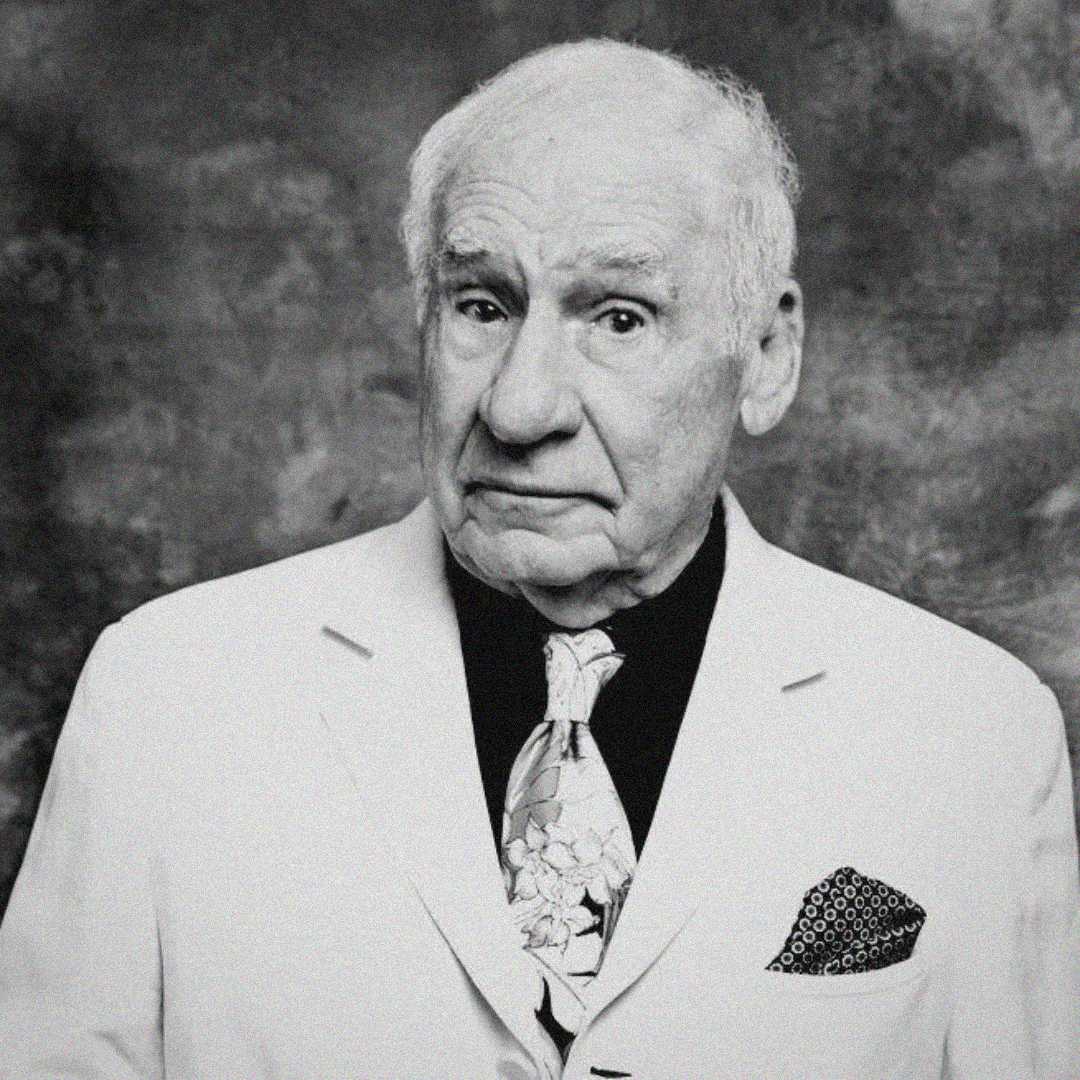 An older man with a slightly raised eyebrow wears a white suit jacket, patterned tie, and pocket square, standing in front of a textured, dark background. The photo is black and white.