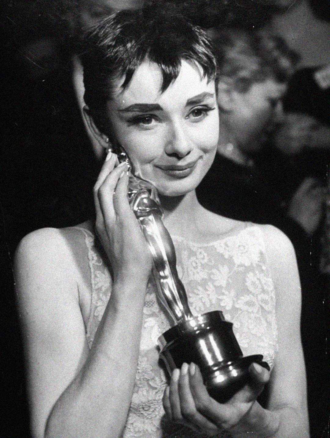 A woman in a light-colored dress holds an Oscar statuette to her face, smiling softly. The image is in black and white, with people blurred in the background.