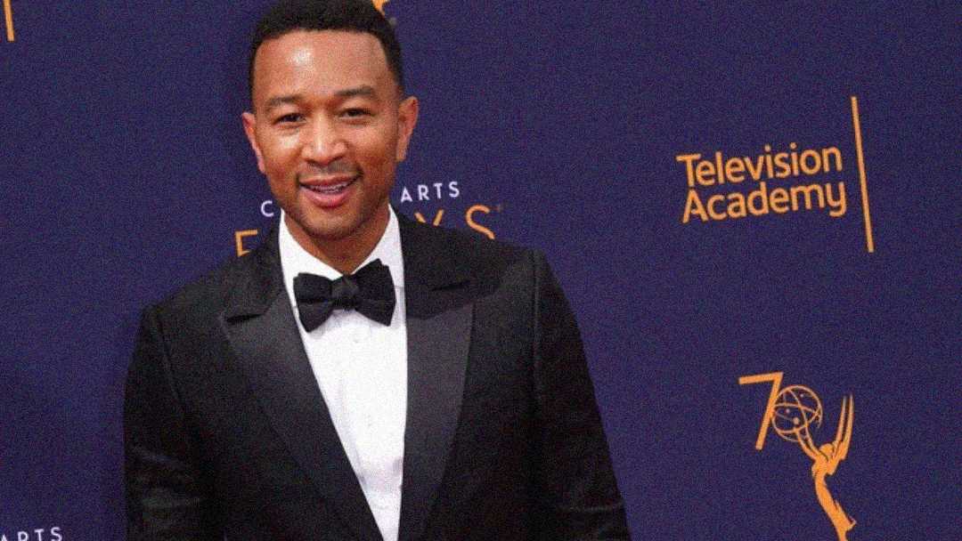 A man in a black tuxedo with a bow tie smiles in front of a blue backdrop with yellow "Television Academy" and Emmy Award logos.