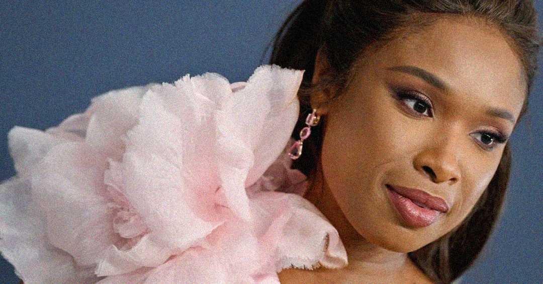 A woman with long, dark hair wears a large, soft pink ruffled garment and dangling pink earrings, set against a dark background. She gazes slightly to the side with a gentle expression.