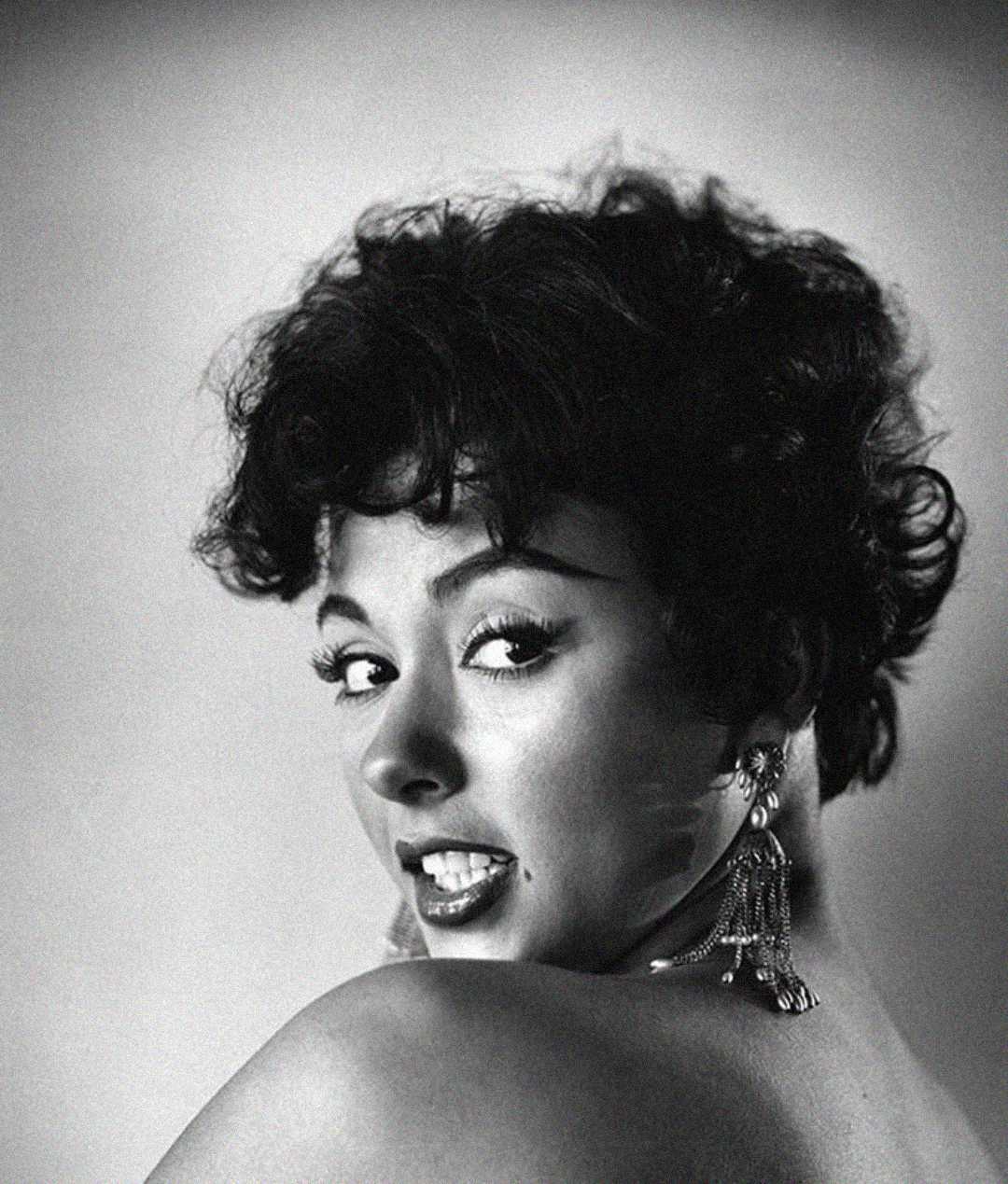 Black and white portrait of a woman with short, curly hair, glancing over her bare shoulder. She wears dramatic eyeliner, dangling earrings, and has an expressive, slightly smiling facial expression.