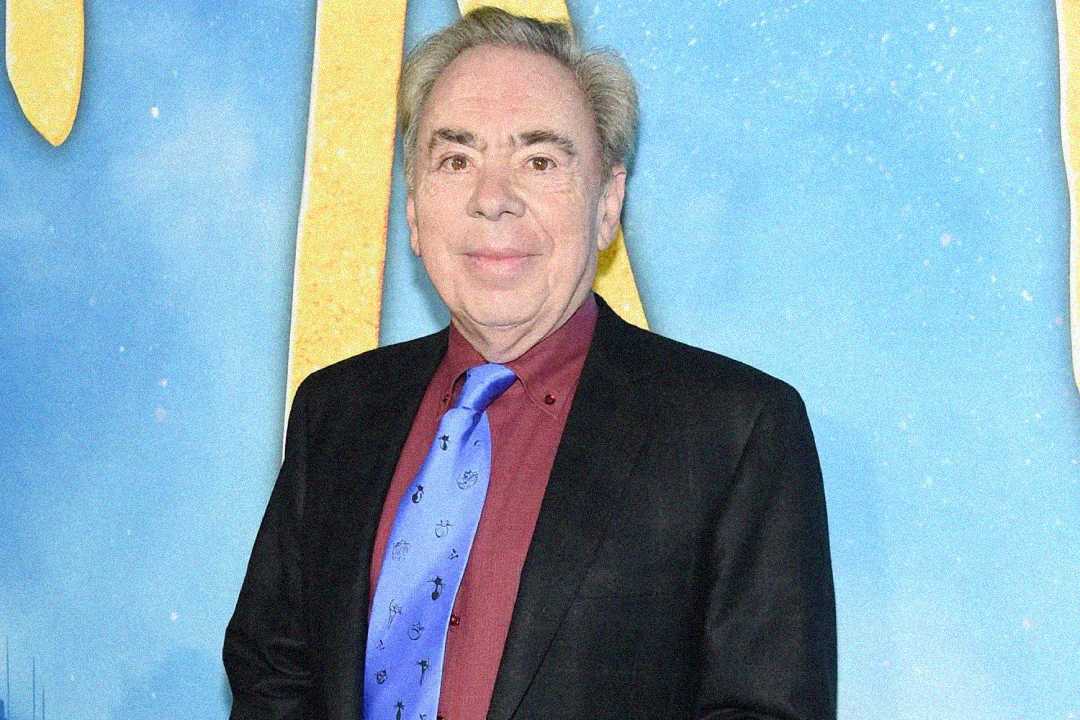 An older man with gray hair wearing a black suit, maroon shirt, and blue tie stands in front of a blue and yellow backdrop, smiling at the camera.