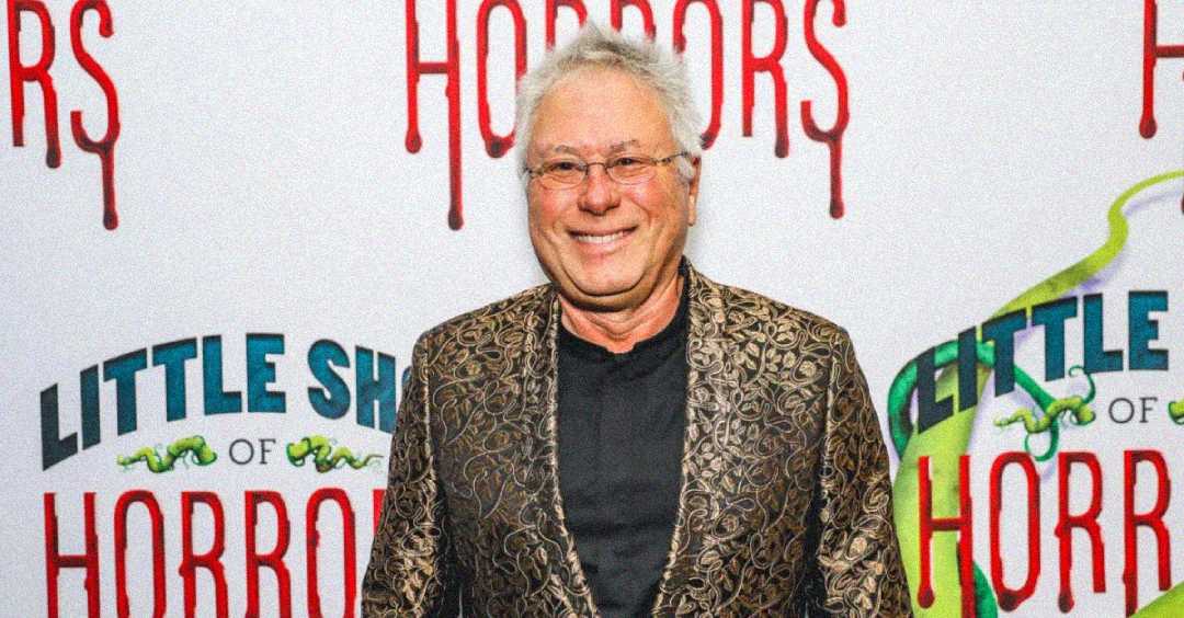 An older man with white hair and glasses, wearing a patterned gold and black jacket, smiles while standing in front of a "Little Shop of Horrors" backdrop.