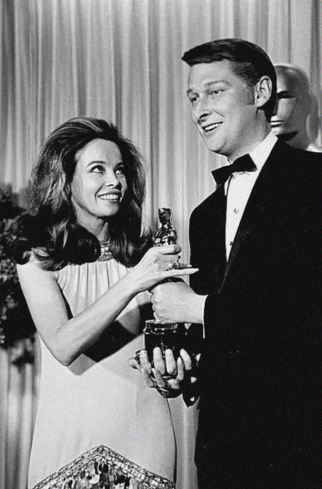 A woman in a dress smiles and points at a man in a tuxedo holding an Oscar statuette, standing together in front of draped curtains at an awards event.