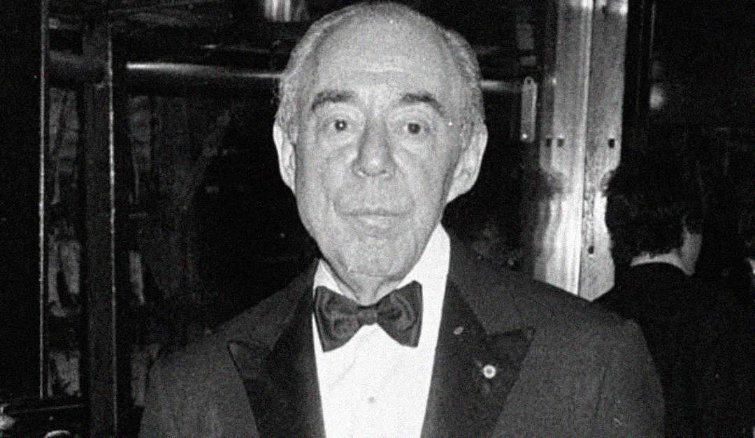 An older man in a tuxedo and bow tie stands indoors, looking at the camera. The image is in black and white, giving it a vintage appearance. The background is blurry, with indistinct people and lights.
