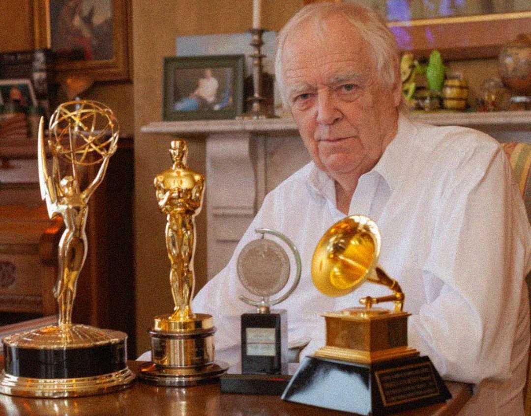 An older man in a white shirt sits at a table displaying four major awards: an Emmy, an Oscar, a Tony, and a Grammy, with framed photos and decorations visible in the background.