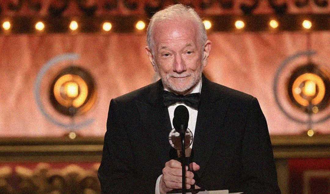 An elderly man in a black tuxedo and bow tie stands at a microphone onstage, smiling and holding a note, with warm lights and decorative circular symbols in the background.