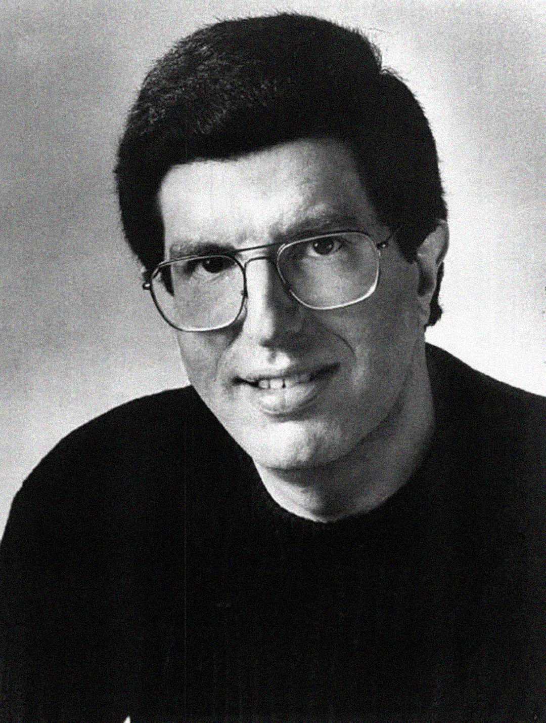 Black and white portrait of a man with short dark hair, wearing large glasses and a dark sweater, smiling slightly at the camera against a plain background.
