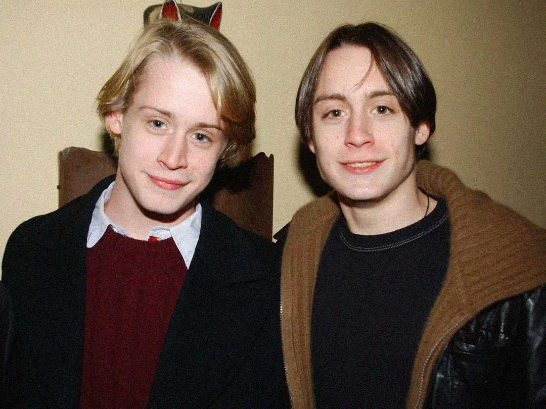 Two young men with light skin are standing closely together and smiling. One has blonde hair and is wearing a collared shirt with a sweater, while the other has brown hair and is wearing a dark shirt with a jacket.
