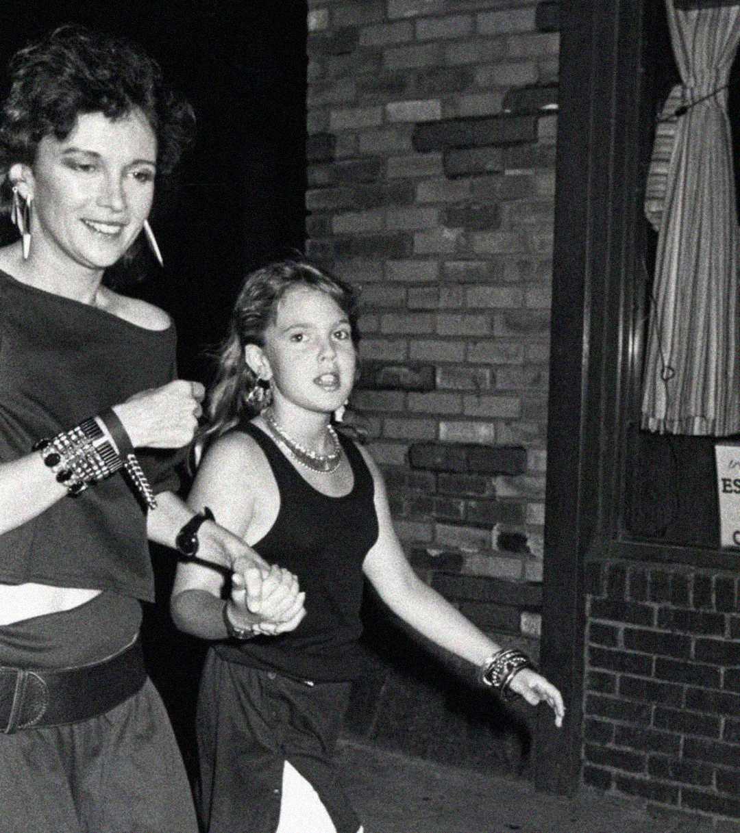 Two people walk hand in hand at night along a sidewalk; one is an adult with short hair and large earrings, and the other is a young girl. Both wear black tops and bracelets, with a brick wall and window in the background.