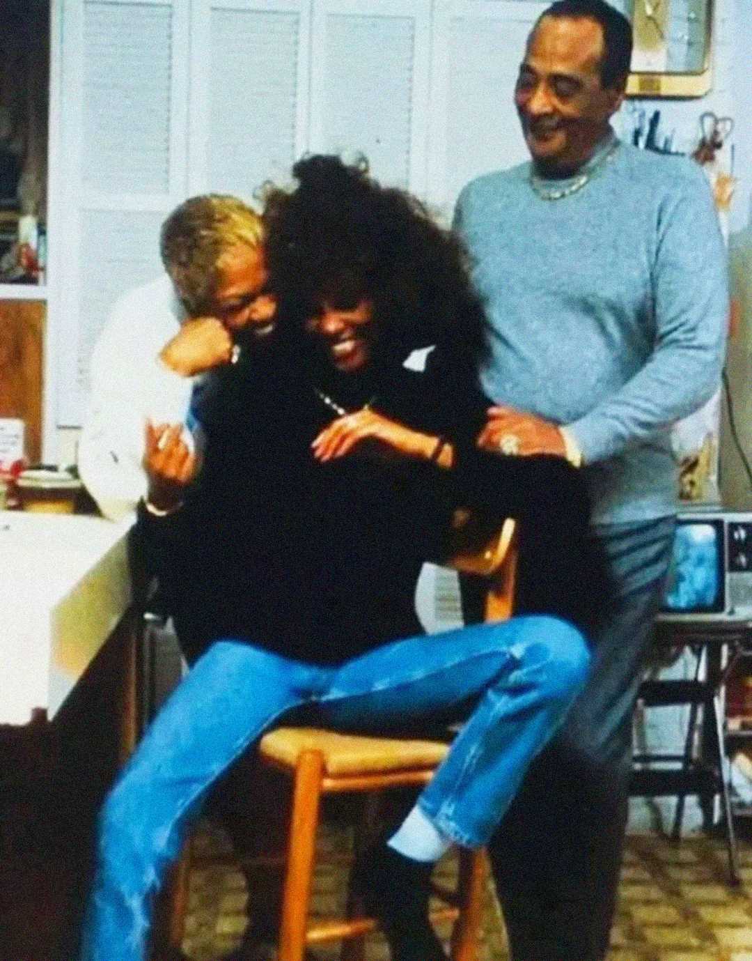 Three people smile and laugh together in a cozy kitchen. One woman sits on a stool with her legs crossed, while the other woman and a man stand close beside her, leaning in affectionately.