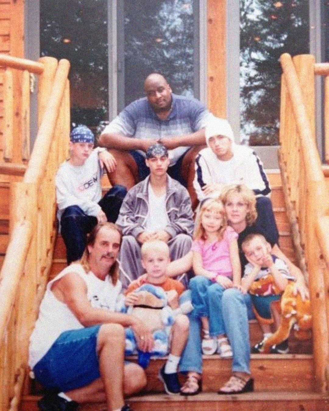 A group of eight people, including adults and children, sit together on wooden steps outside a log cabin. Some wear bandanas and casual clothing, and a few children hold stuffed toys. Trees are visible through the glass door behind them.