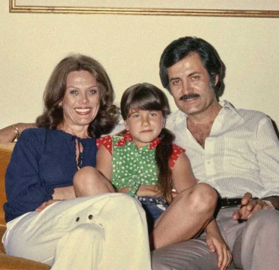 A woman, a young girl, and a man sit close together on a couch, smiling for the camera in a warmly lit room. The woman and man have their arms around the girl, suggesting a family portrait.
