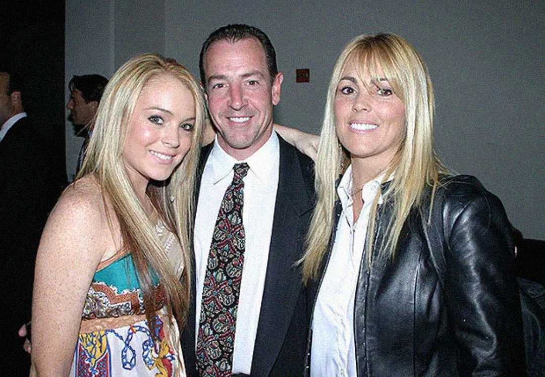 Three people pose together indoors, smiling at the camera. The young woman on the left wears a colorful patterned dress, the man in the center wears a suit and patterned tie, and the woman on the right wears a black jacket and white shirt.